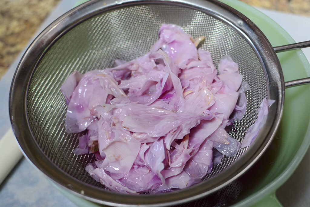 Spent, faded purple peony petals being strained in a metal mesh colander over a green bowl.