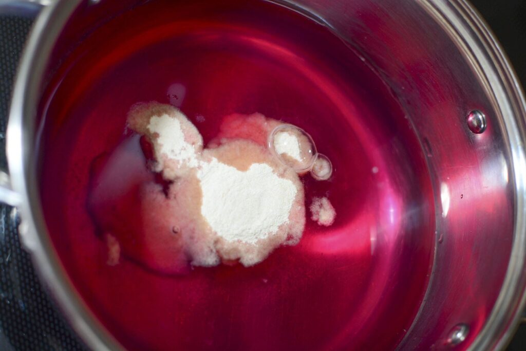 White powdered pectin sitting on top of a deep pink liquid in a stainless steel pot before being mixed.