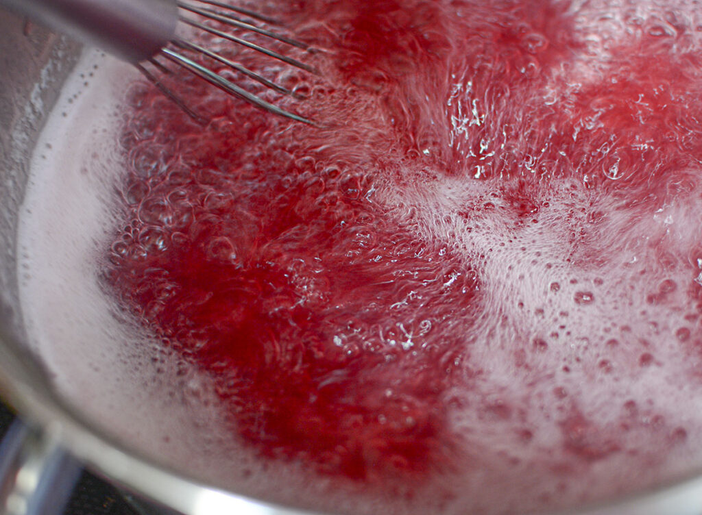 A metal whisk stirring a boiling, bubbling mixture of bright red peony liquid in a stainless steel pot.