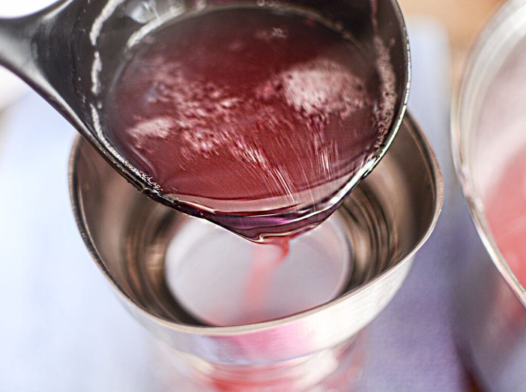 Liquid pink peony liquid being ladled from a pot through a metal funnel into a glass jar.