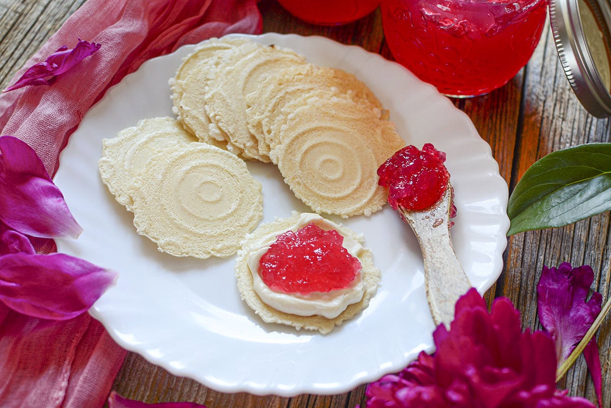 A white plate featuring thin, round crackers with cream cheese and a dollop of bright pink peony jelly, with a jar of jelly in the background.