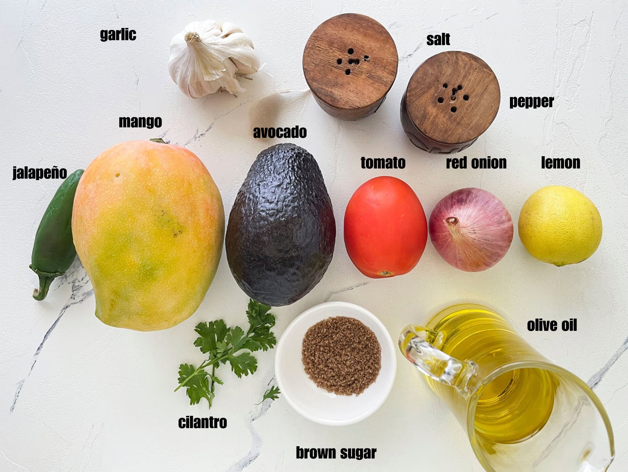 a flat lay arrangement of raw ingredients, including a mango, an avocado, a tomato, an onion, a lemon, garlic, herbs, oil, and spices on a white counter.