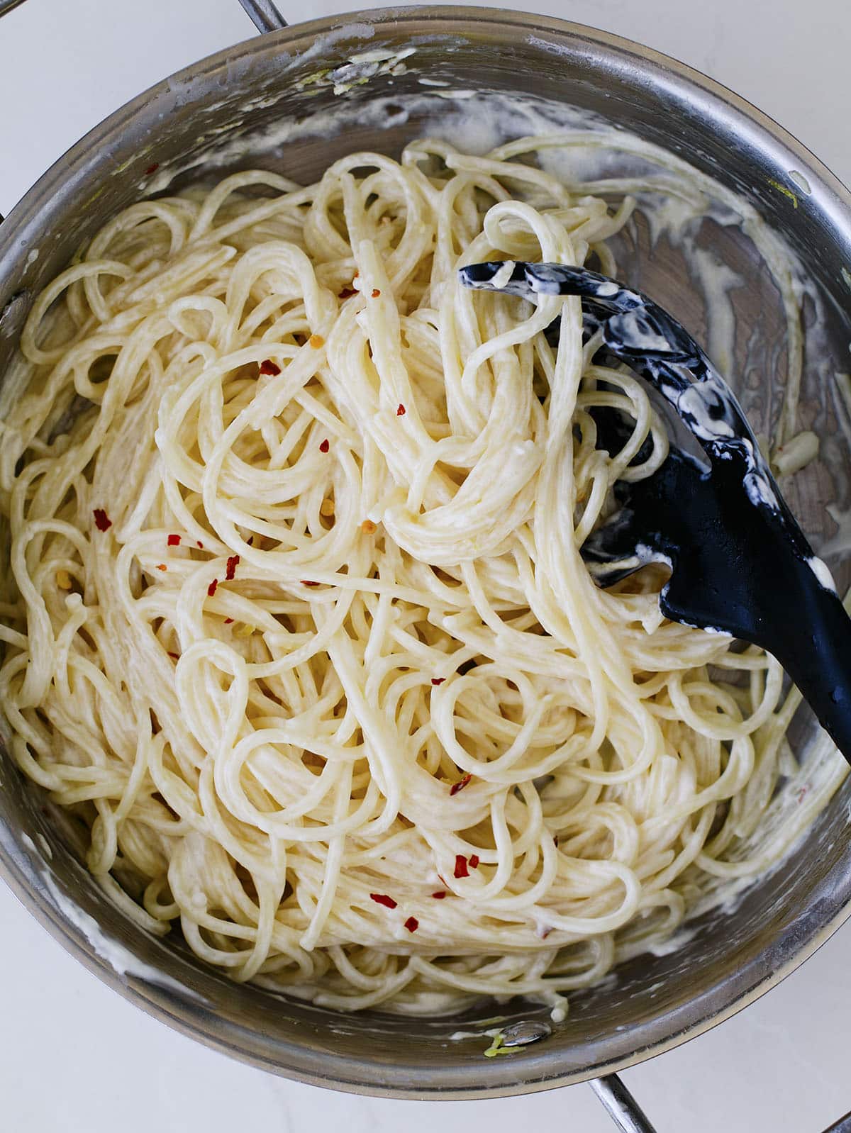 Creamy pasta noodles being tossed with red pepper flakes in a stainless steel pan.