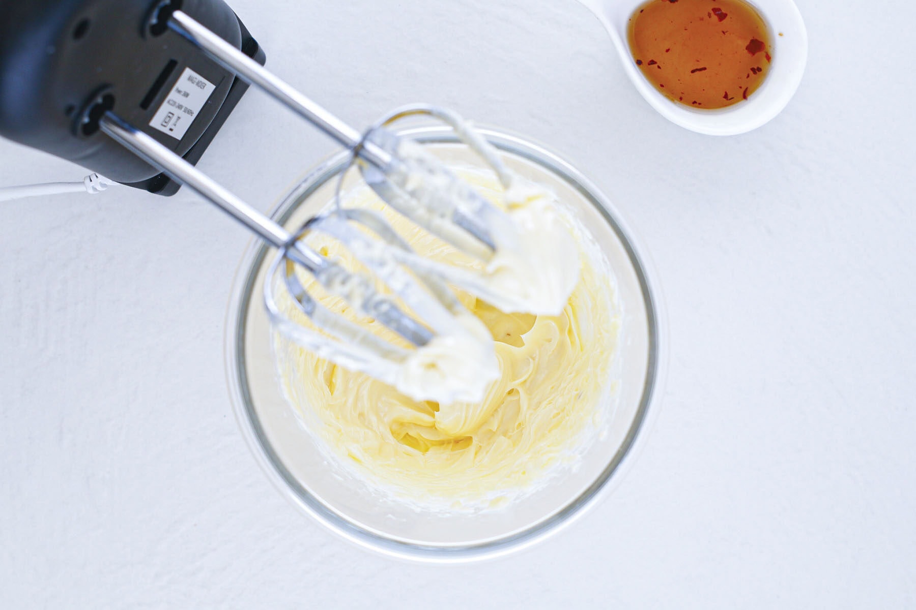 An electric hand mixer blending a creamy pale yellow mixture in a glass bowl.