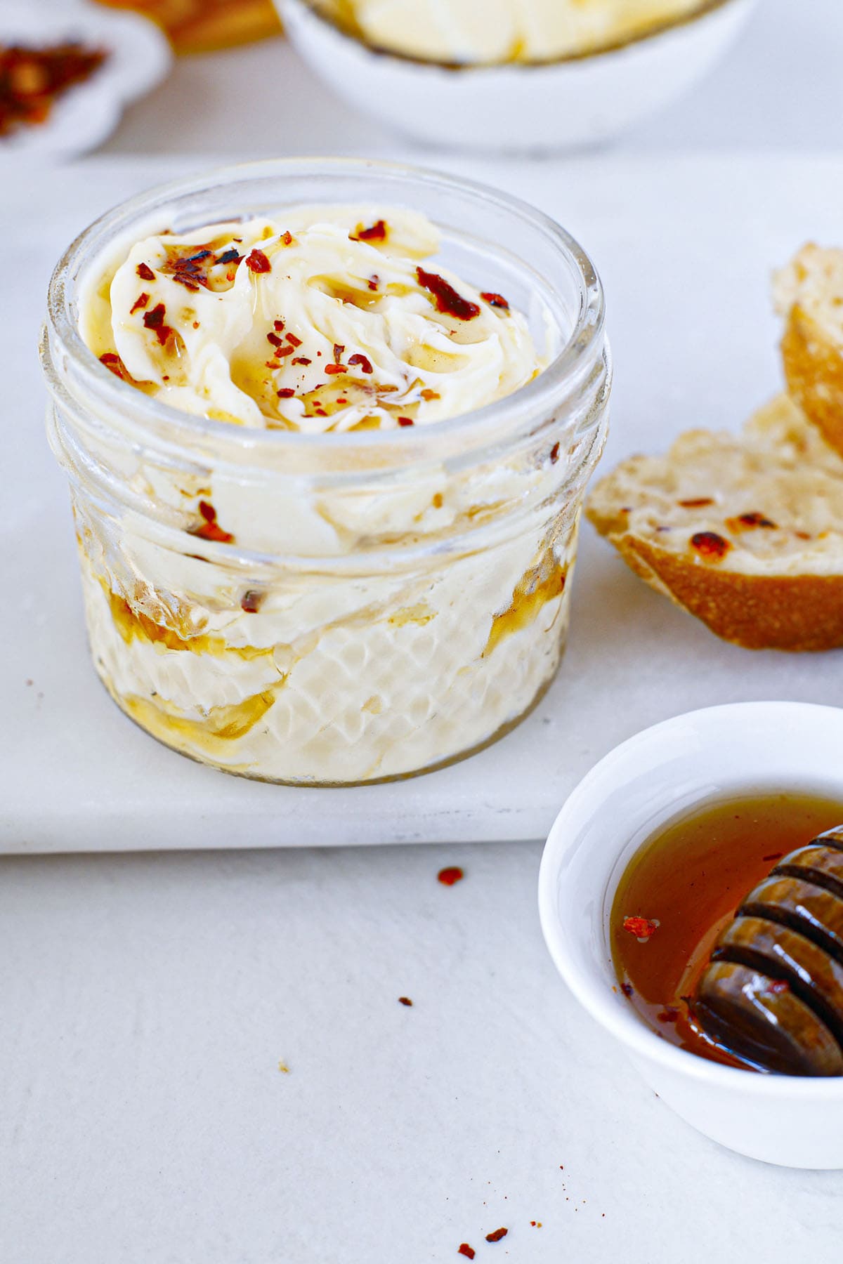 Whipped honey spread in a small jar next to a piece of bread and a small bowl of honey.