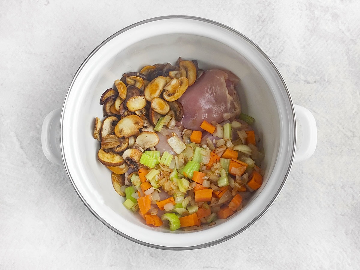 Raw chicken thighs, sautéed mushrooms, and chopped vegetables combined in a soup pot.