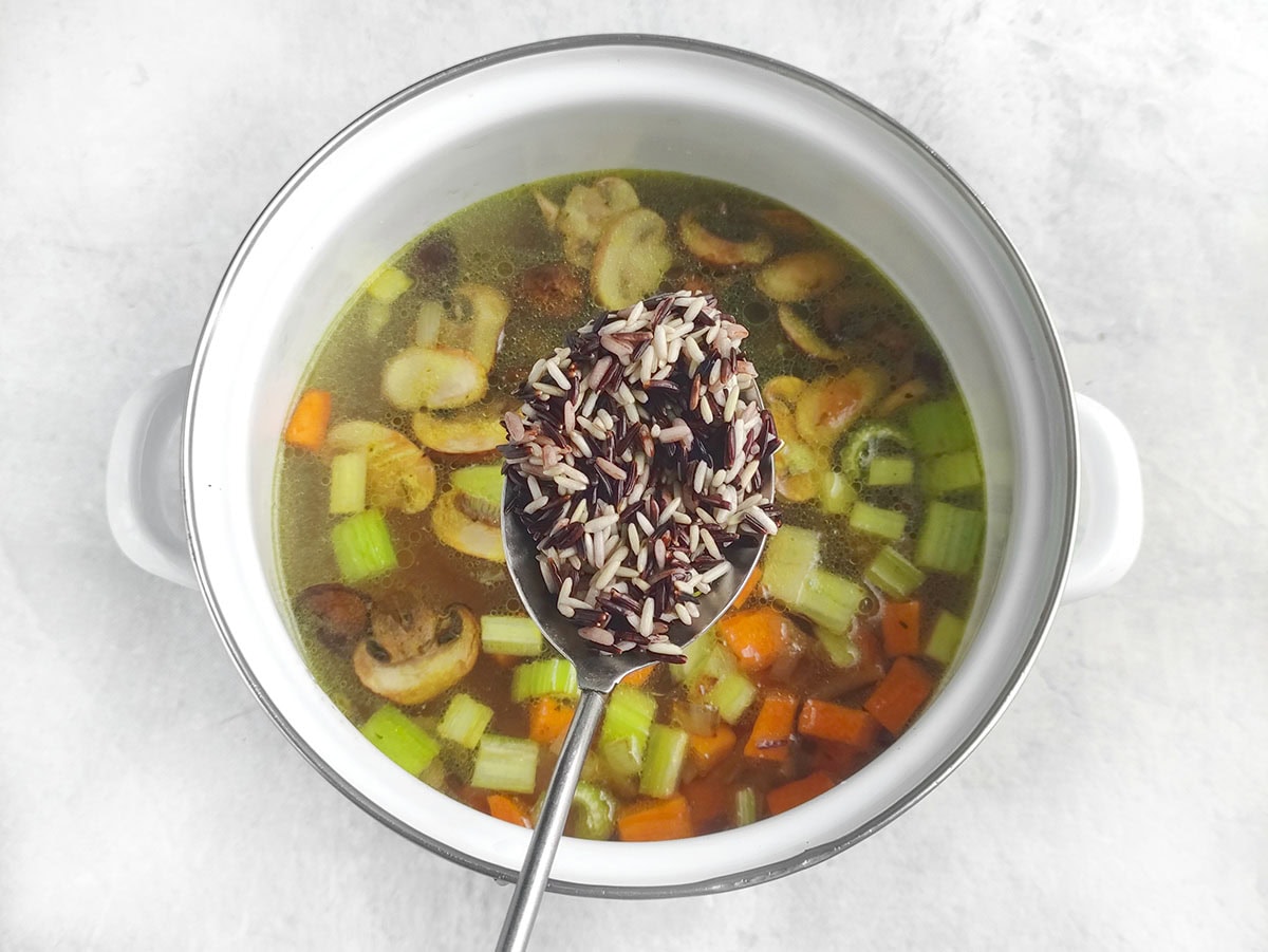 A spoonful of wild rice being added to a pot of vegetable and mushroom broth.