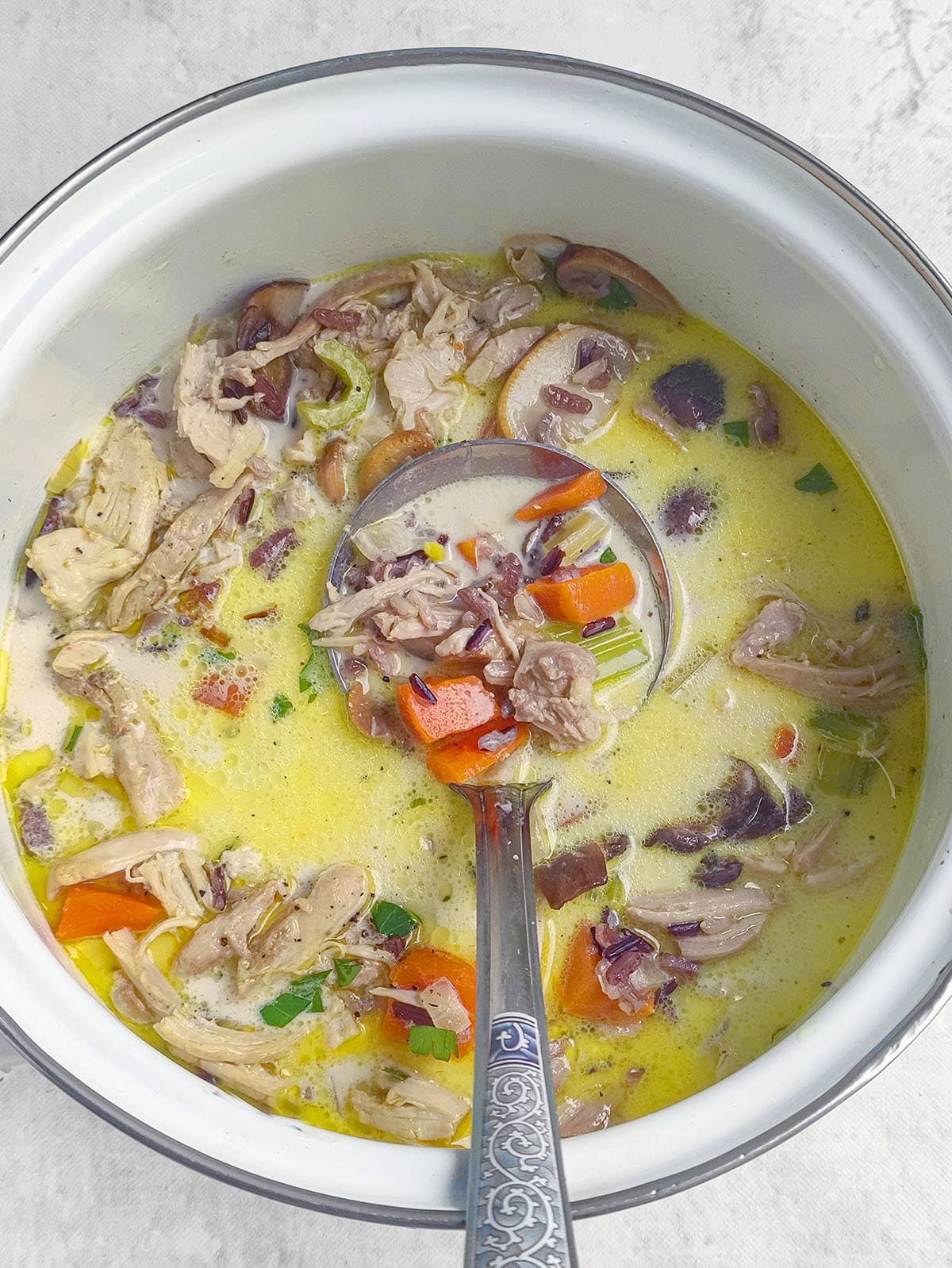 A ladle scooping up creamy chicken, mushroom, carrot, and wild rice soup from a pot.