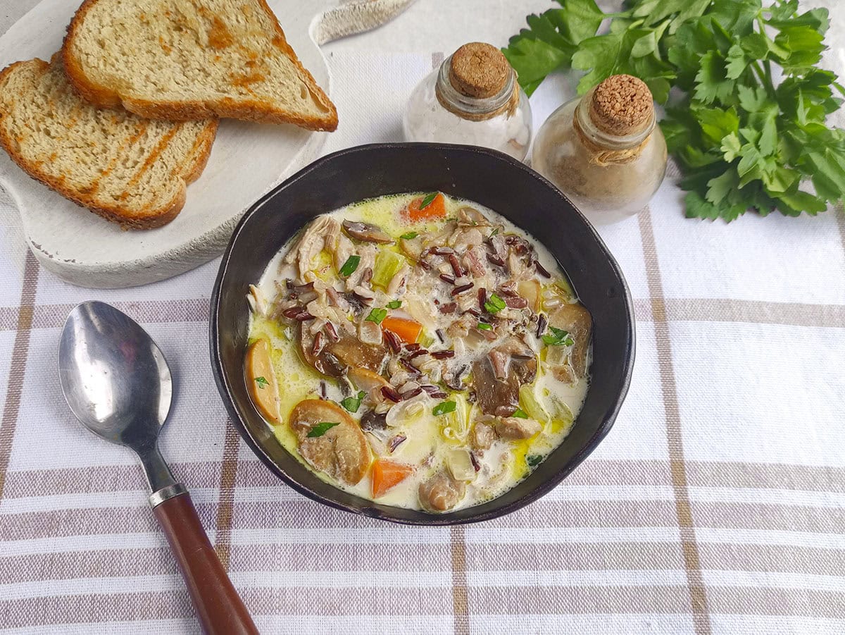 A bowl of chicken, mushroom, and wild rice soup served on a plaid cloth next to sliced bread.
