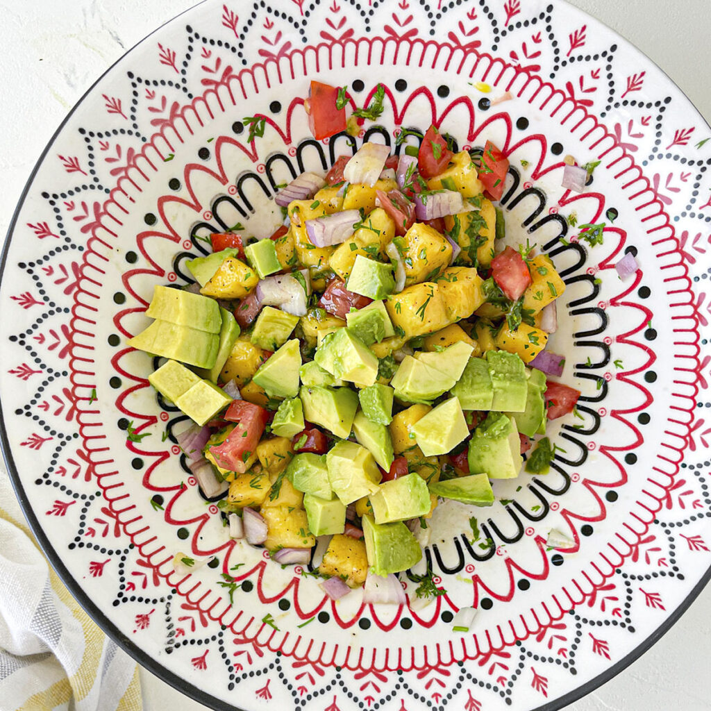 Diced avocado layered on top of a mixed fruit and vegetable salad in a patterned bowl.