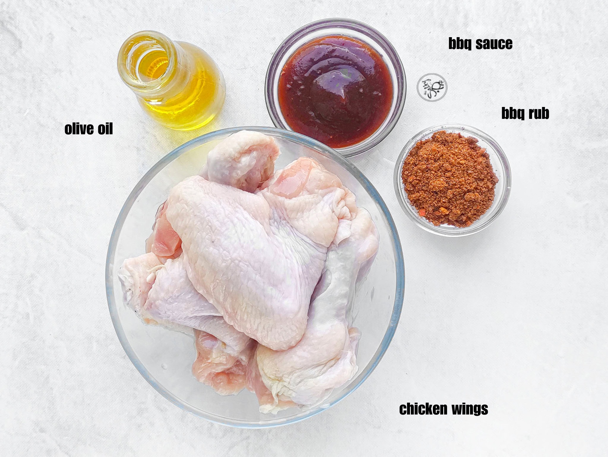  Raw chicken wings in a glass bowl placed alongside oil, barbecue sauce, and spice rub in small containers.