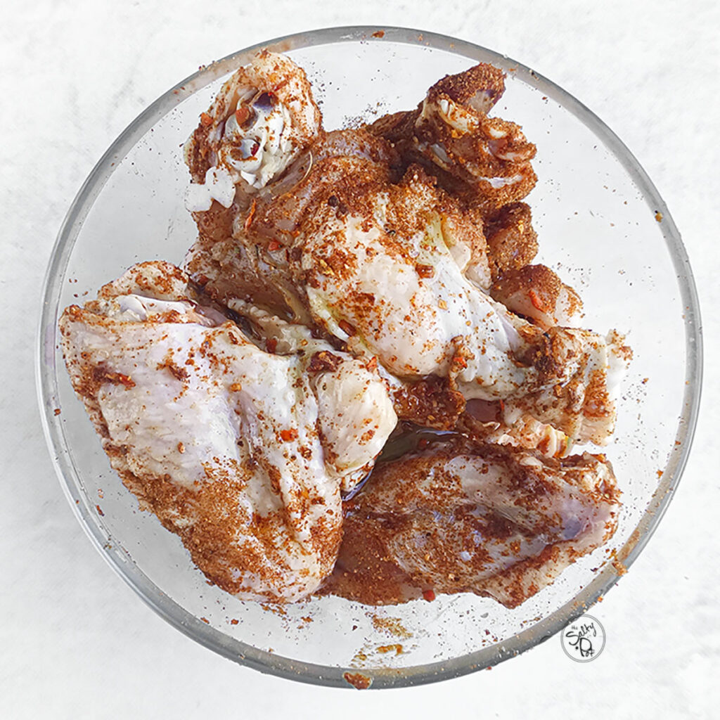 Raw chicken wings seasoned with a dry spice rub in a clear mixing bowl.
