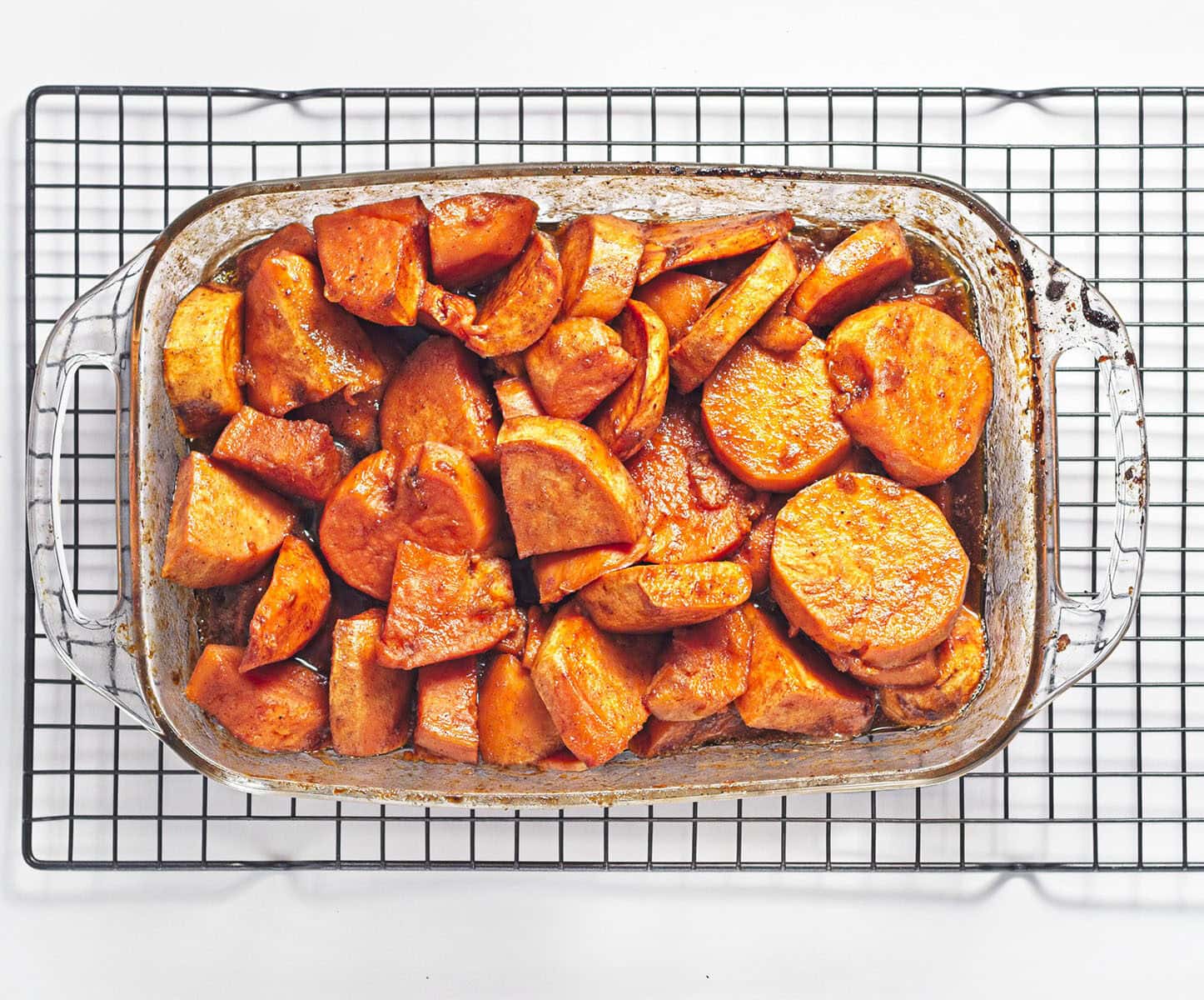 A glass baking pan filled with sliced sweet potatoes resting on a wire cooling rack.
