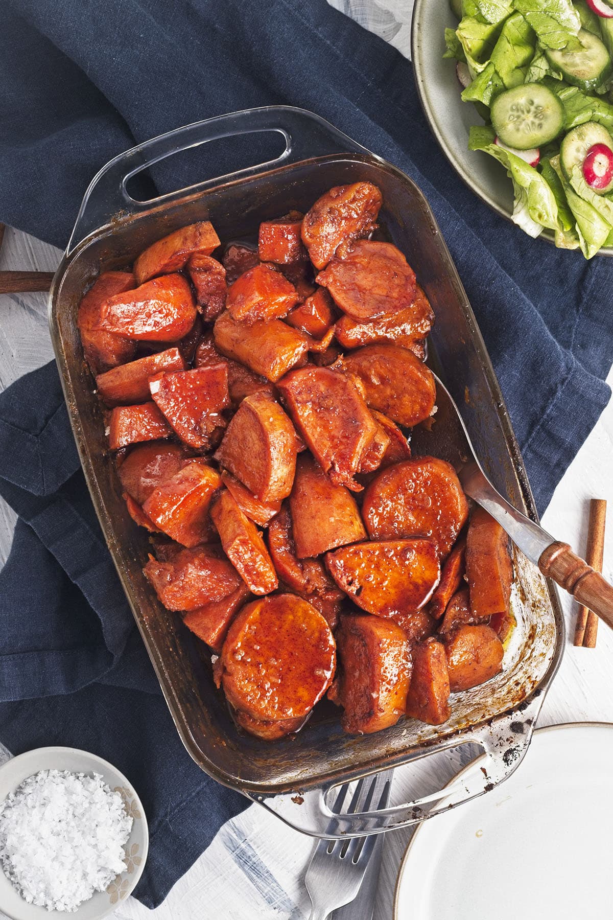 Overhead view of glazed sweet potatoes in a baking dish, served alongside side salads and salt.
