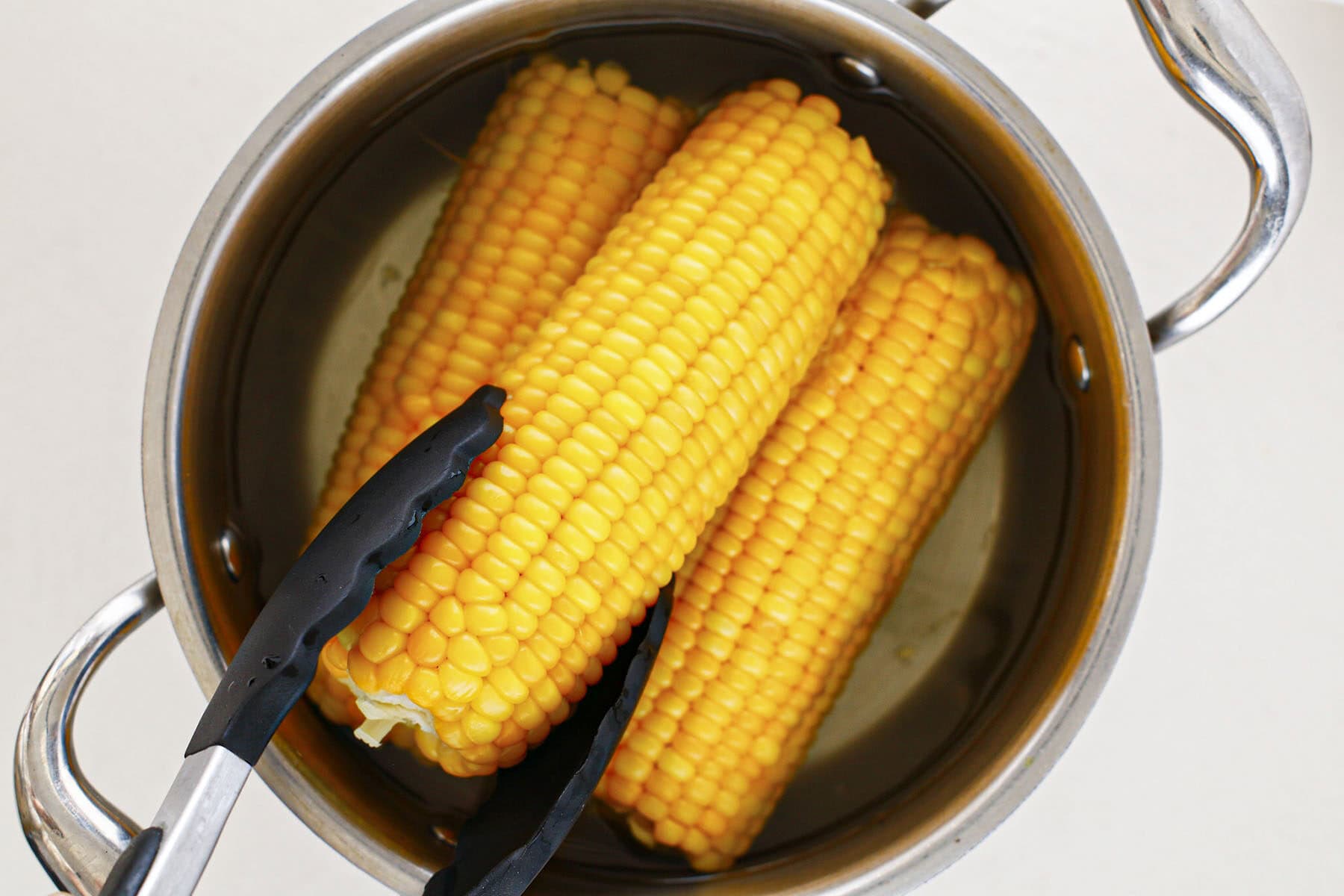 Black tongs lifting boiled corn on the cob from a stainless steel pot of water