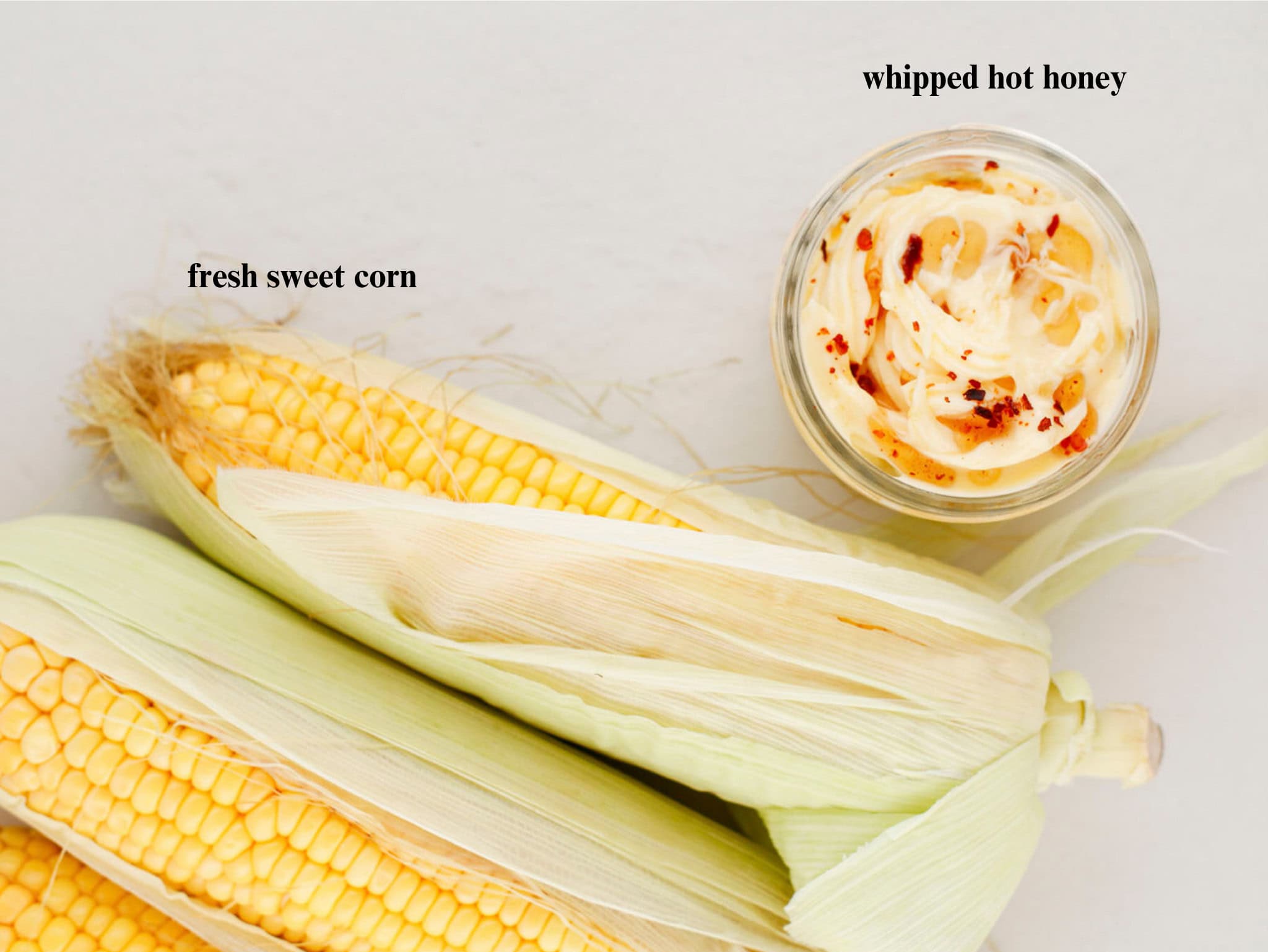 Fresh ears of corn in husks next to a glass jar of whipped honey butter topped with chili flakes
