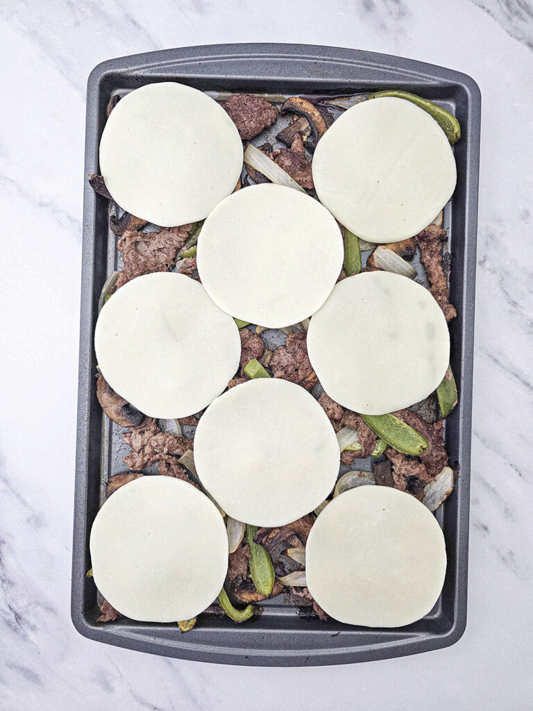 Round slices of cheese placed over cooked meat and vegetables on a sheet pan.