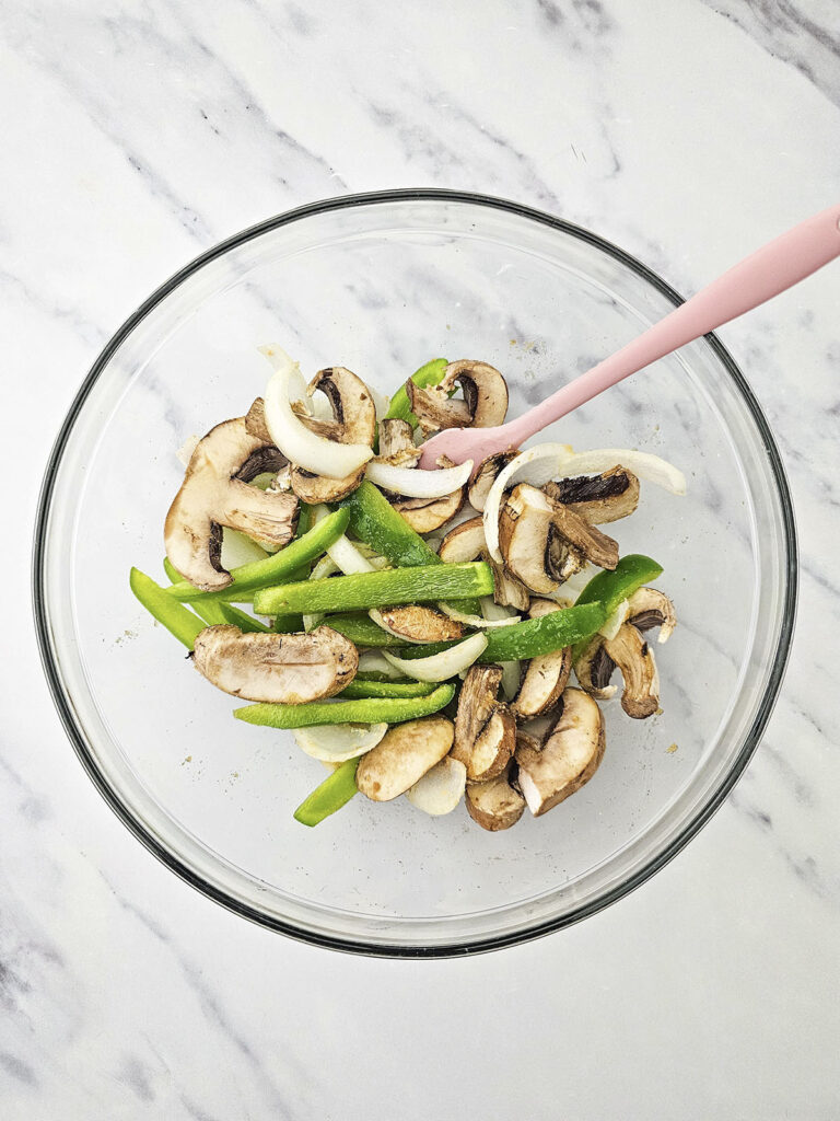  Sliced mushrooms, green peppers, and onions mixed in a glass bowl.
