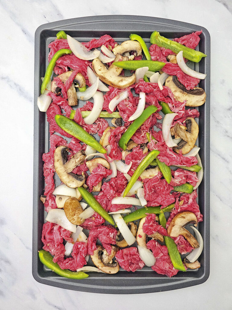 Uncooked beef, sliced peppers, mushrooms, and onions arranged on a baking sheet.