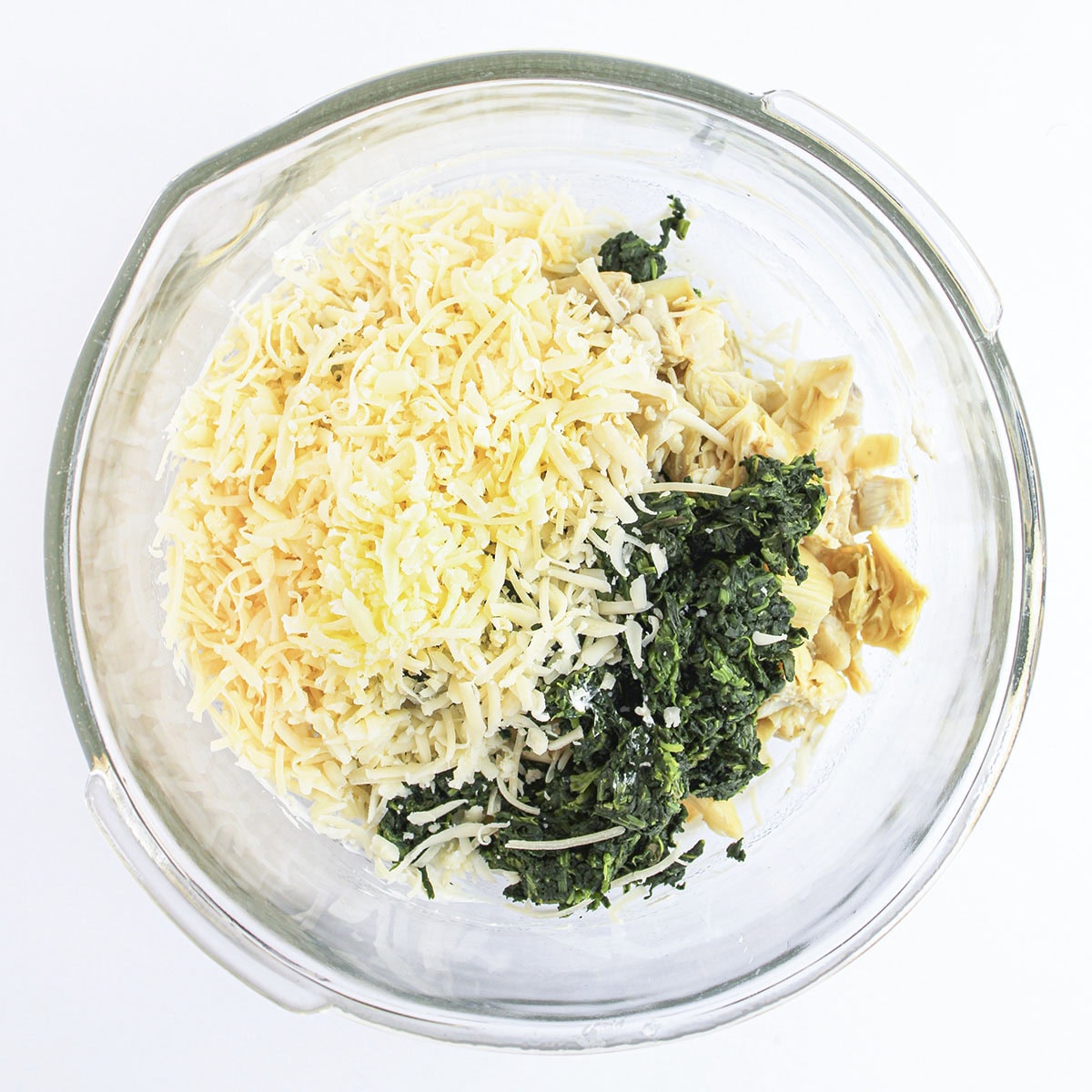 Chopped spinach, artichoke hearts, and shredded cheese separated in a clear mixing bowl