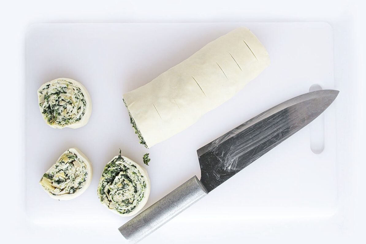 Rolled pastry log being sliced into pinwheels with a chef's knife on a white cutting board