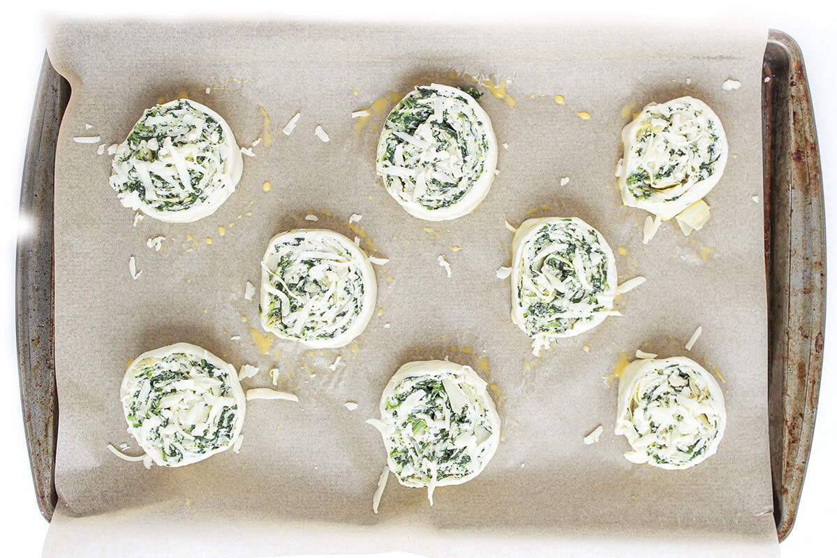 Eight unbaked, sliced spinach artichoke pinwheels arranged on a parchment-lined baking sheet