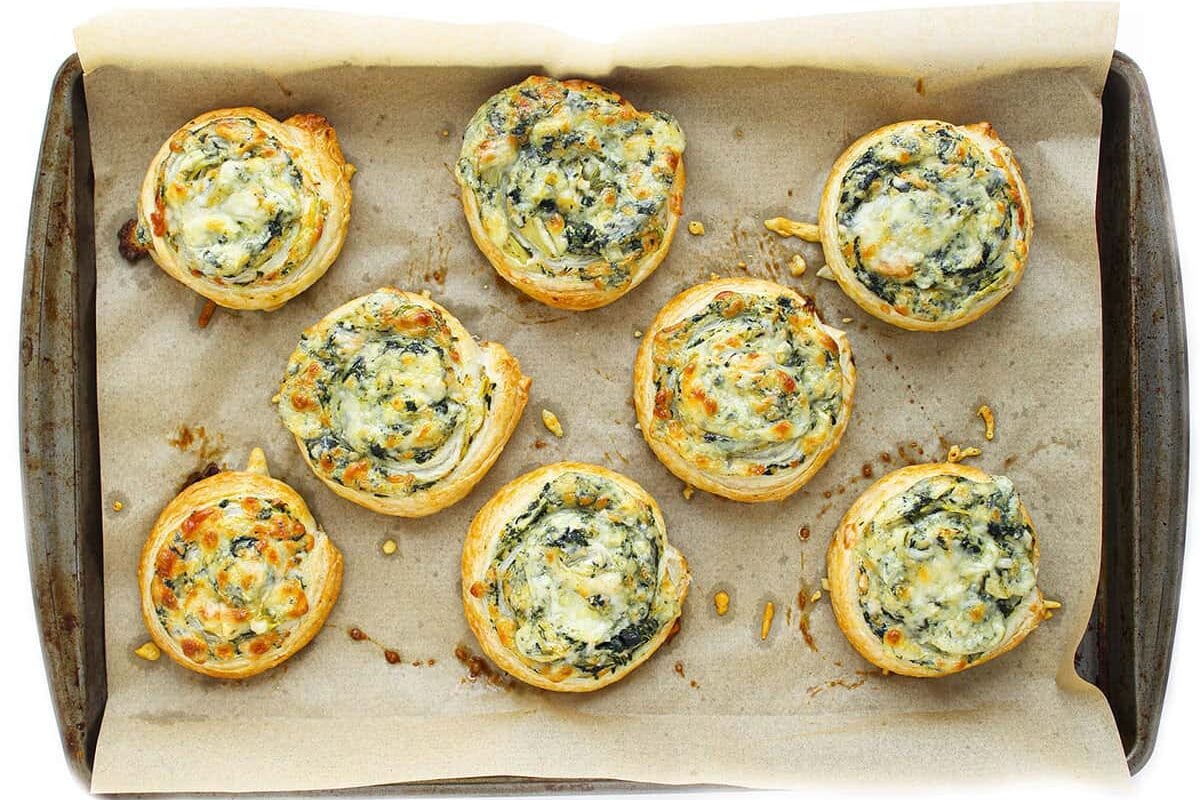 Top-down view of eight baked spinach artichoke pinwheels on a parchment-lined baking sheet