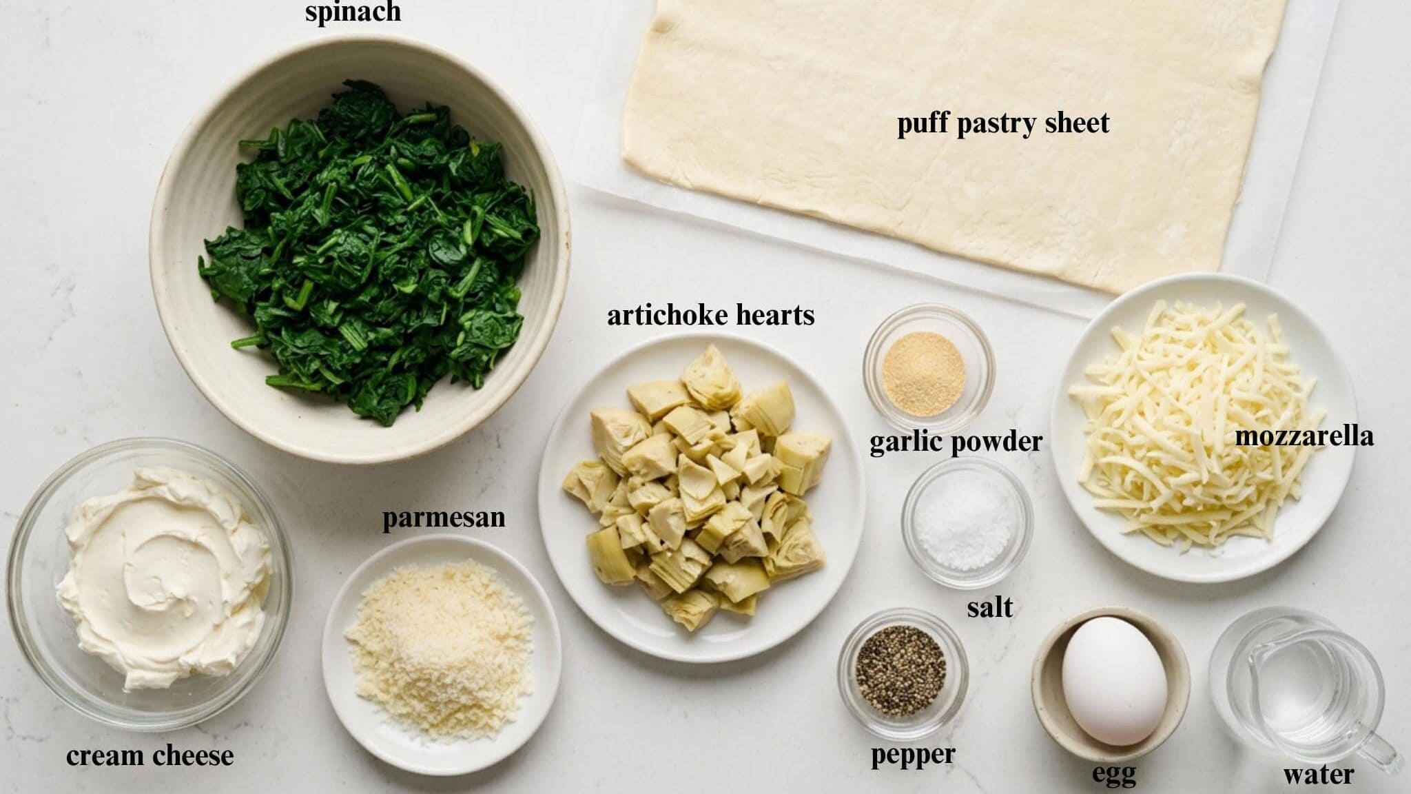 Prepped ingredients for spinach artichoke pinwheels arranged on a white counter including puff pastry, spinach, artichokes, cheeses, and egg