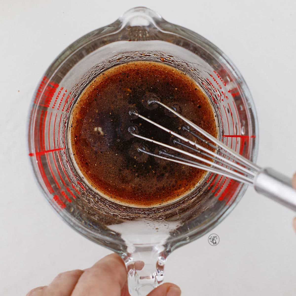 Hand holding a small whisk in a glass measuring cup filled with the dressing.