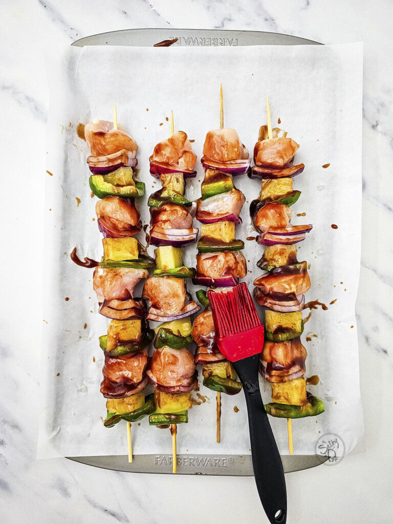 Four unbaked skewers lined up on parchment paper being brushed with teriyaki glaze