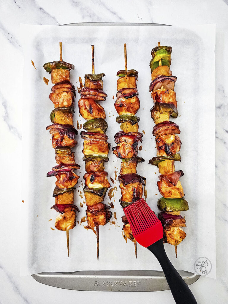 Four cooked chicken kabobs on parchment paper being brushed with sauce