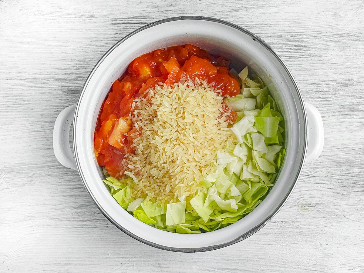 Uncooked rice, chopped cabbage, and tomato sauce added into a pot.