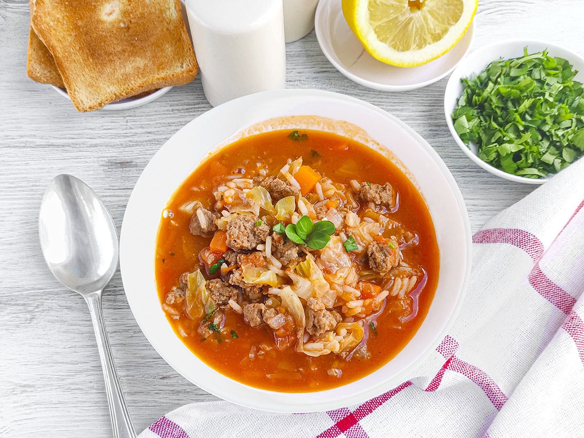 White bowl filled with hearty meat and vegetable soup served next to toast slices and herbs.
