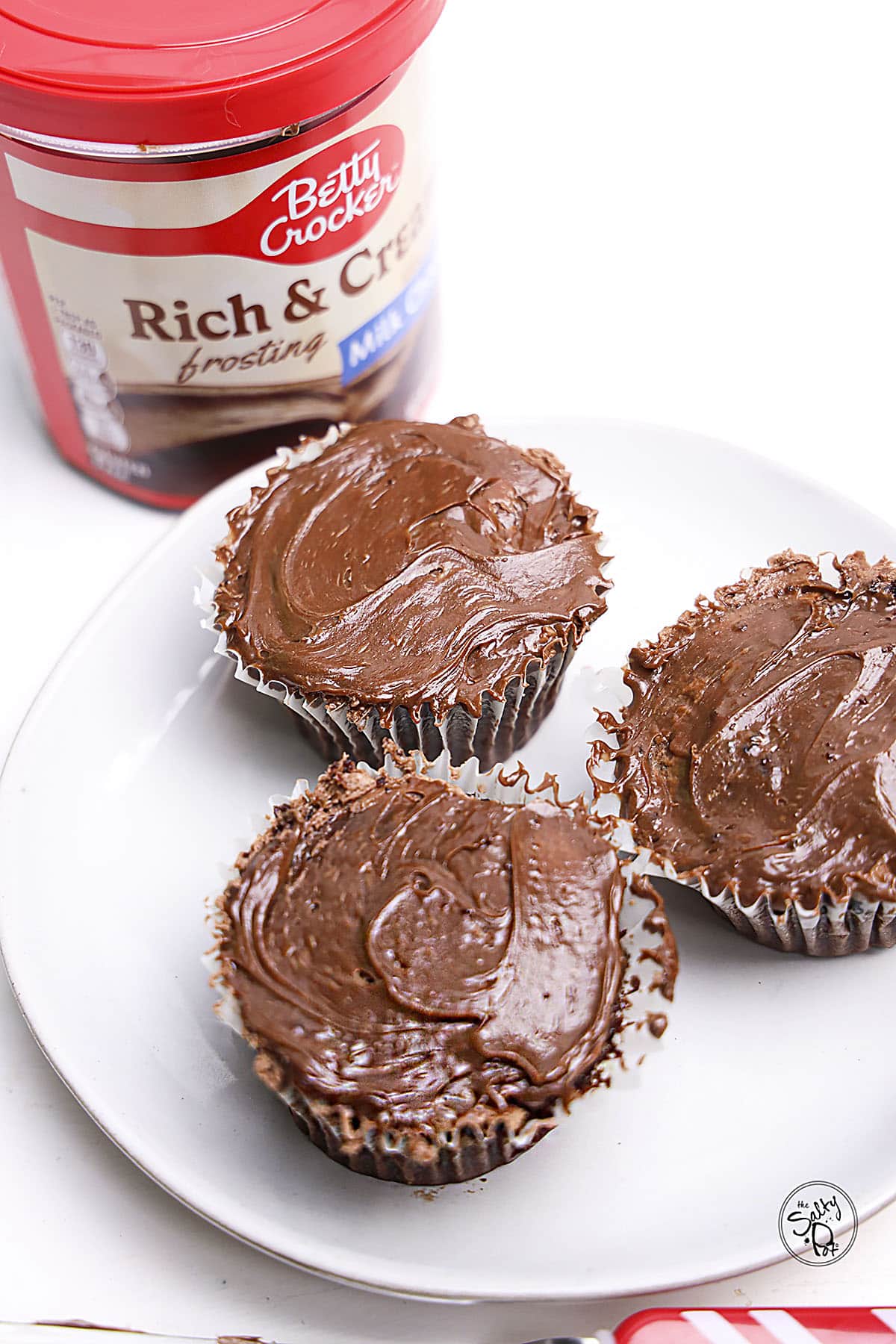 Three freshly frosted chocolate cupcakes on a white plate next to a container of chocolate frosting.