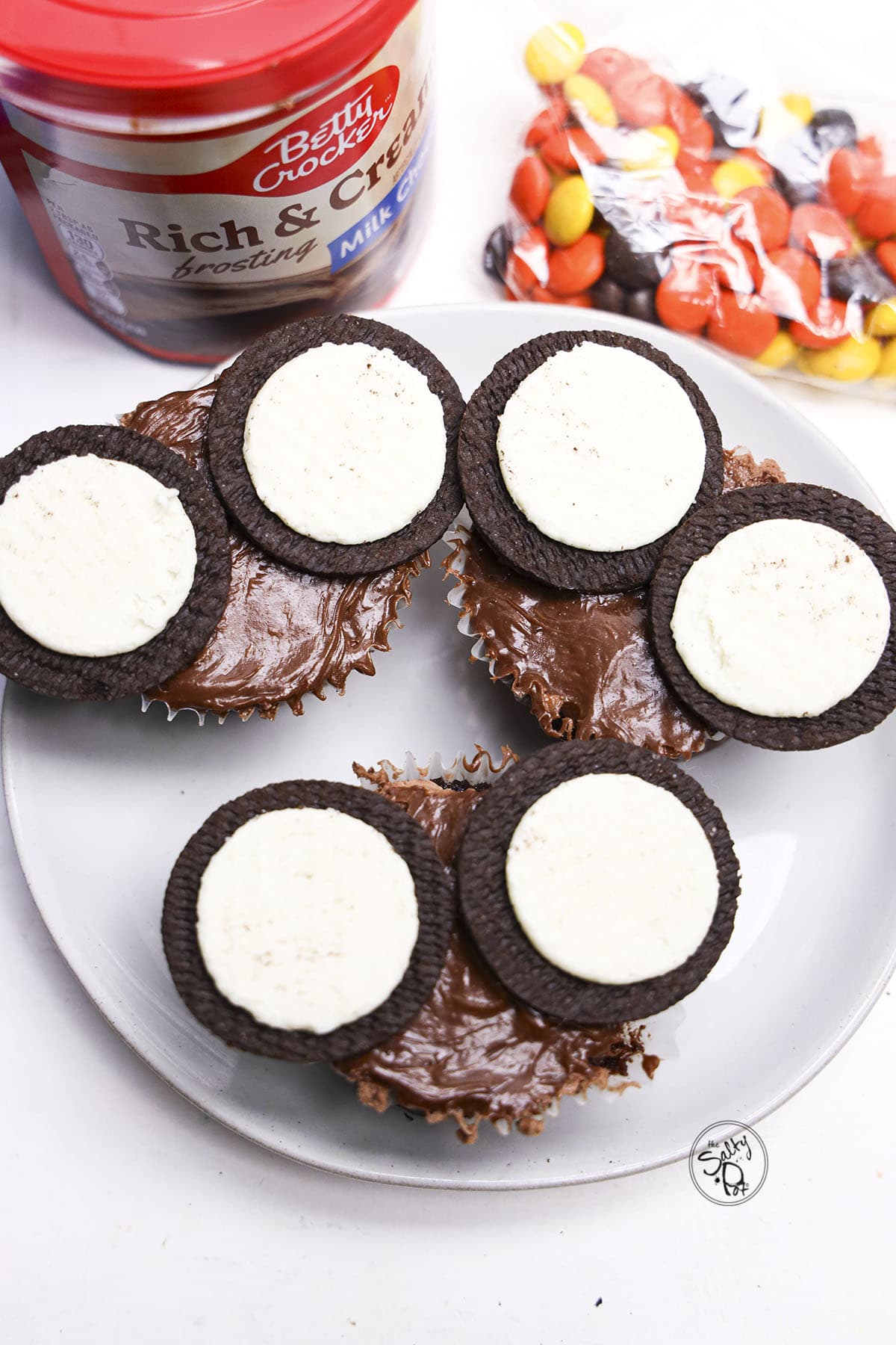 Frosted chocolate cupcakes topped with open cream-filled cookies, positioned next to a tub of frosting and candies