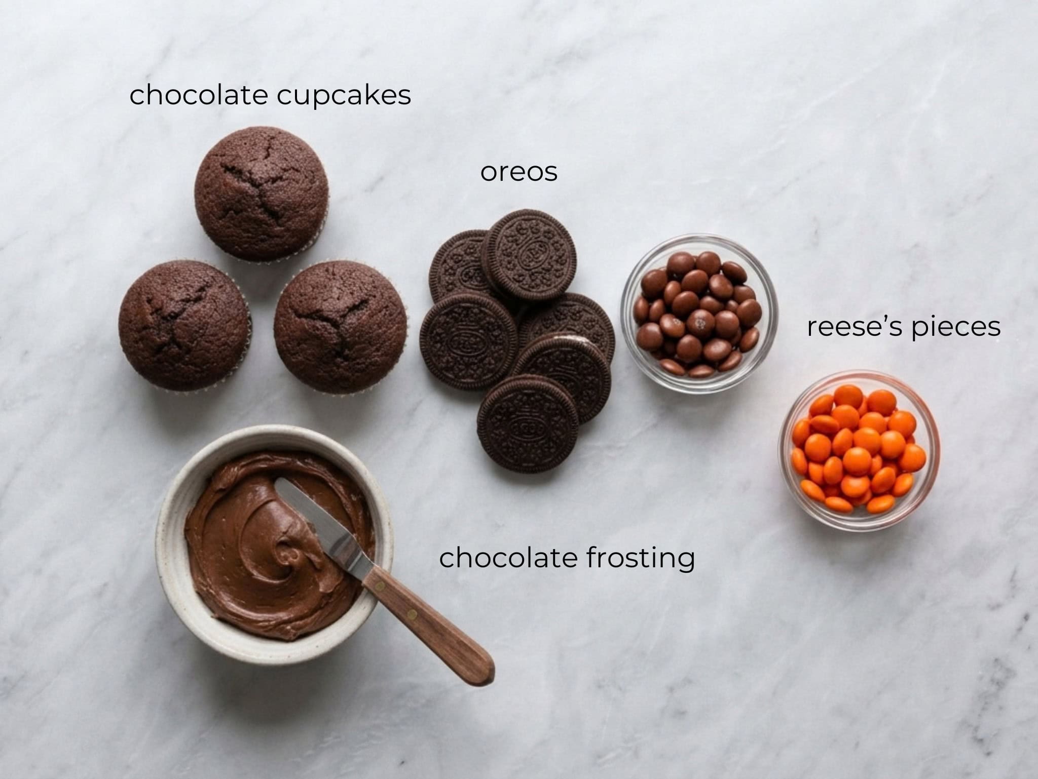 Prepped dessert ingredients including chocolate cupcakes, cookies, frosting, and candies laid out on a marble surface.