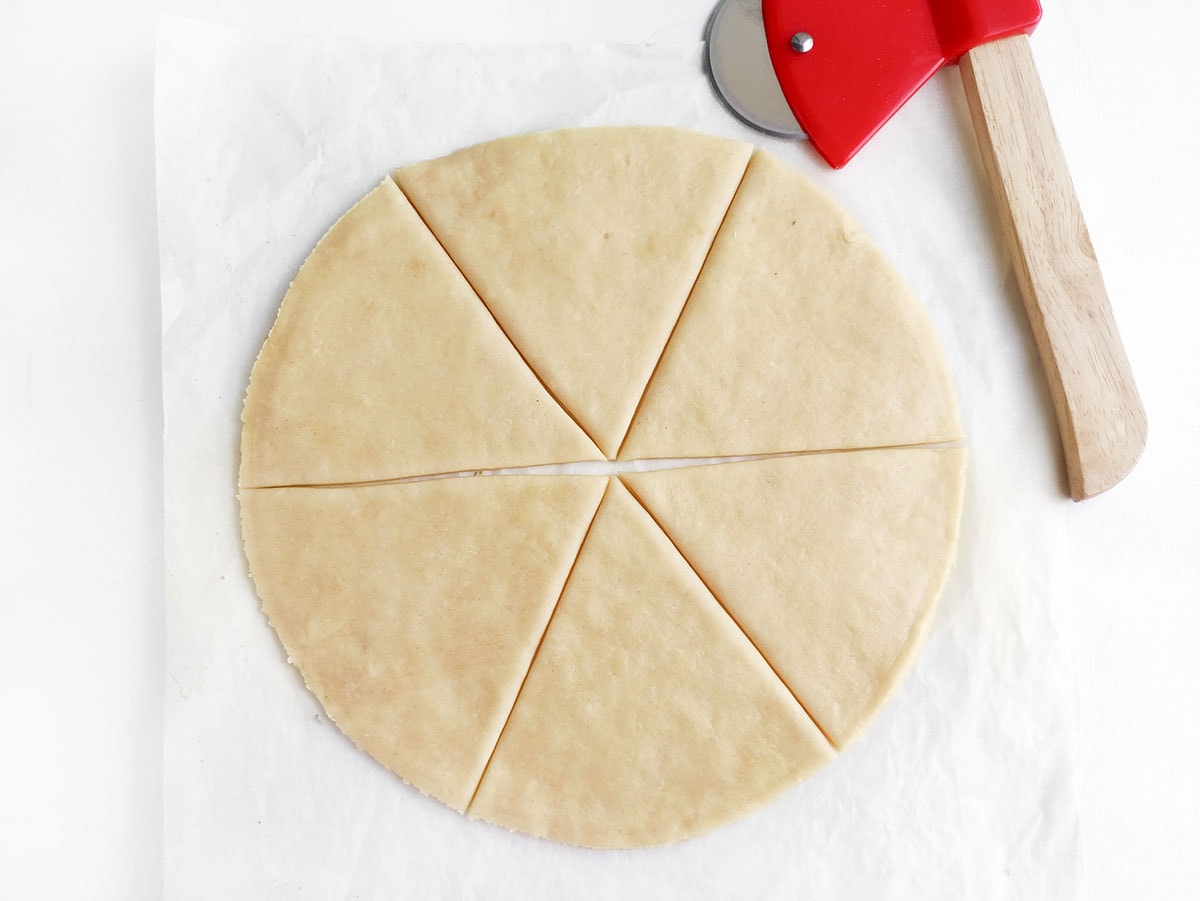 Round sheet of pastry dough sliced into six triangular wedges next to a pastry cutter
