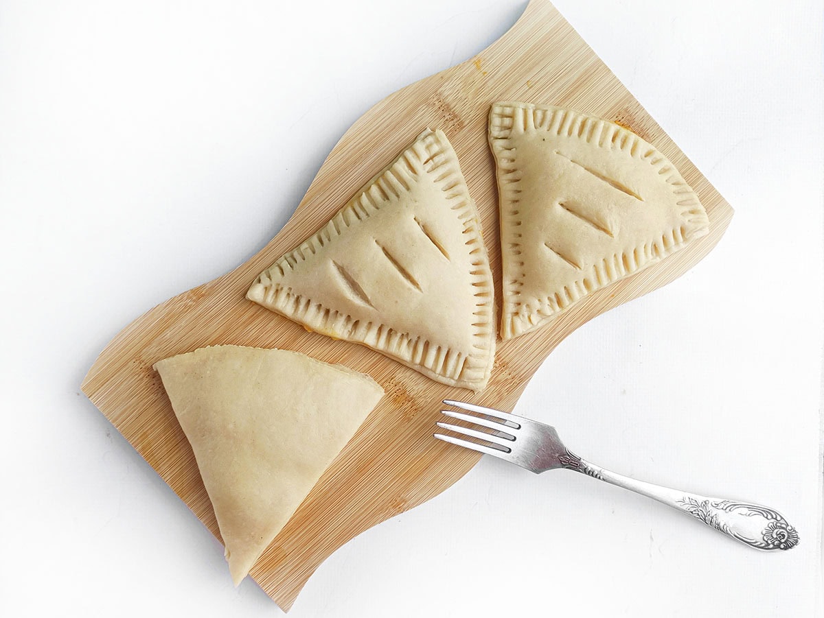 assembled unbaked pastry triangles with crimped edges sitting on a wooden board next to a fork